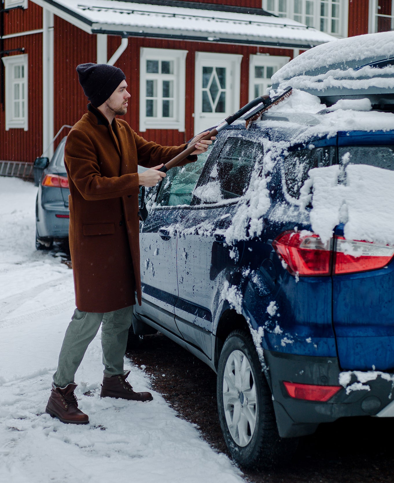 A man cleaning his car from snow in front of a house.