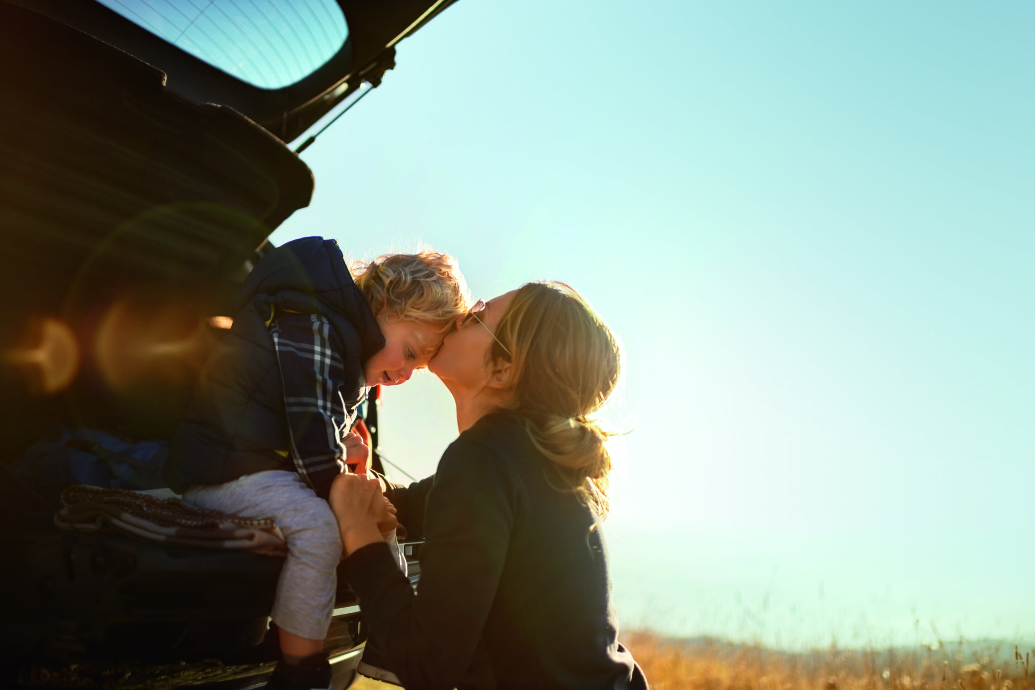 614643666 Mother and little son travel by car in mountains.Little boy sitting in car trunk while they makes a break and mother gives him a kiss.
