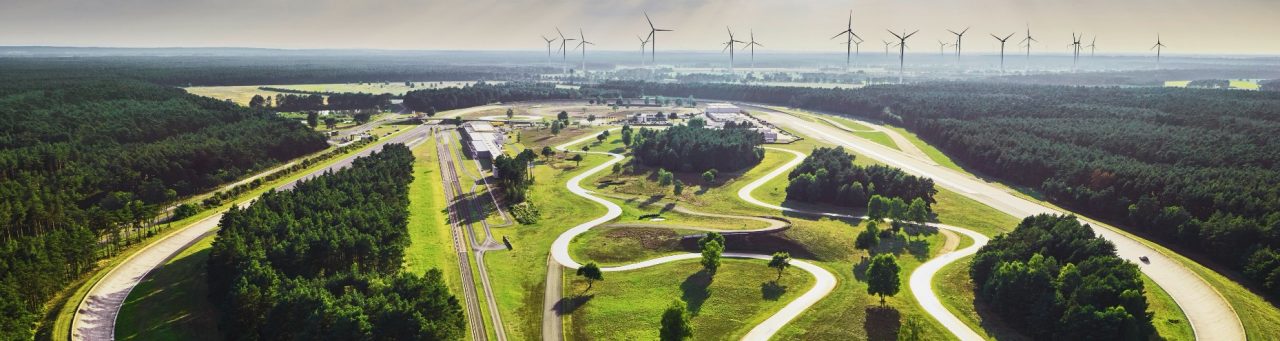 Vista aerea di una grande pista di prova per autoveicoli circondata da una foresta, con strade tortuose, tratti rettilinei, edifici e turbine eoliche sullo sfondo.